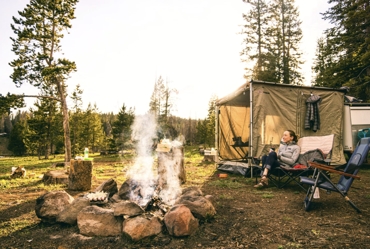 woman-lounging-on-camping-chair-in-front-of-bonfire-in-sunny-forest-with-square-tent-behind-her